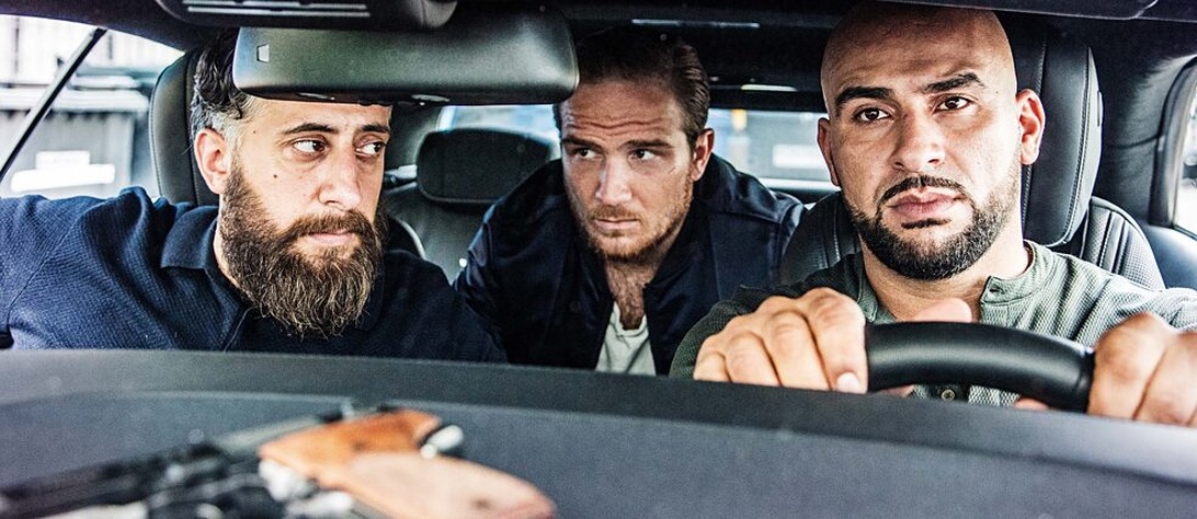 Drei Männer im Innenraum eines Autos mit ernstem, intensivem Gesichtsausdruck. Das Bild hat eine filmische Qualität mit kontrastreicher Beleuchtung. Zwei Männer tragen Bärte, der dritte sitzt auf dem Rücksitz. Dies scheint entweder ein Filmstill, eine Musikvideoszene oder ein Werbefoto für ein Musik- oder Filmprojekt zu sein. Im Vordergrund ist etwas sichtbar, das wie eine Waffe aussieht.